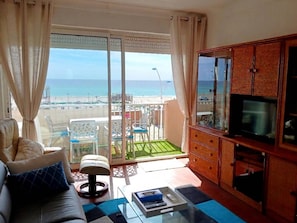 A living room with a sofa, a coffee table, a TV, and a balcony with a view of the beach and ocean.