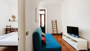 Ein kompakter Wohnraum mit einer Couch, einem Fernseher auf einem weißen Schrank, einer Holzleiter und einem kleinen Tisch mit einer Vase.