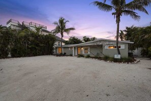 A two-story house with a front porch, surrounded by palm trees and a sandy driveway.