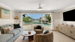 Amplia sala de estar con un sofá gris, una mesa de centro redonda y un sillón color beige. Un televisor grande está montado en la pared, y se ve una piscina y vegetación al exterior.