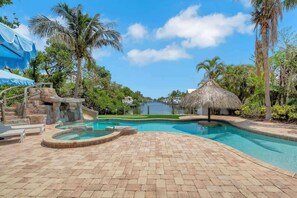 A swimming pool with a hot tub, palm trees, and a thatched-roof hut.