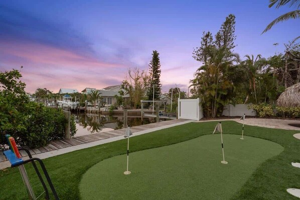 A well-maintained artificial green lawn with a putting green and a small pond, surrounded by tropical vegetation and a wooden deck.