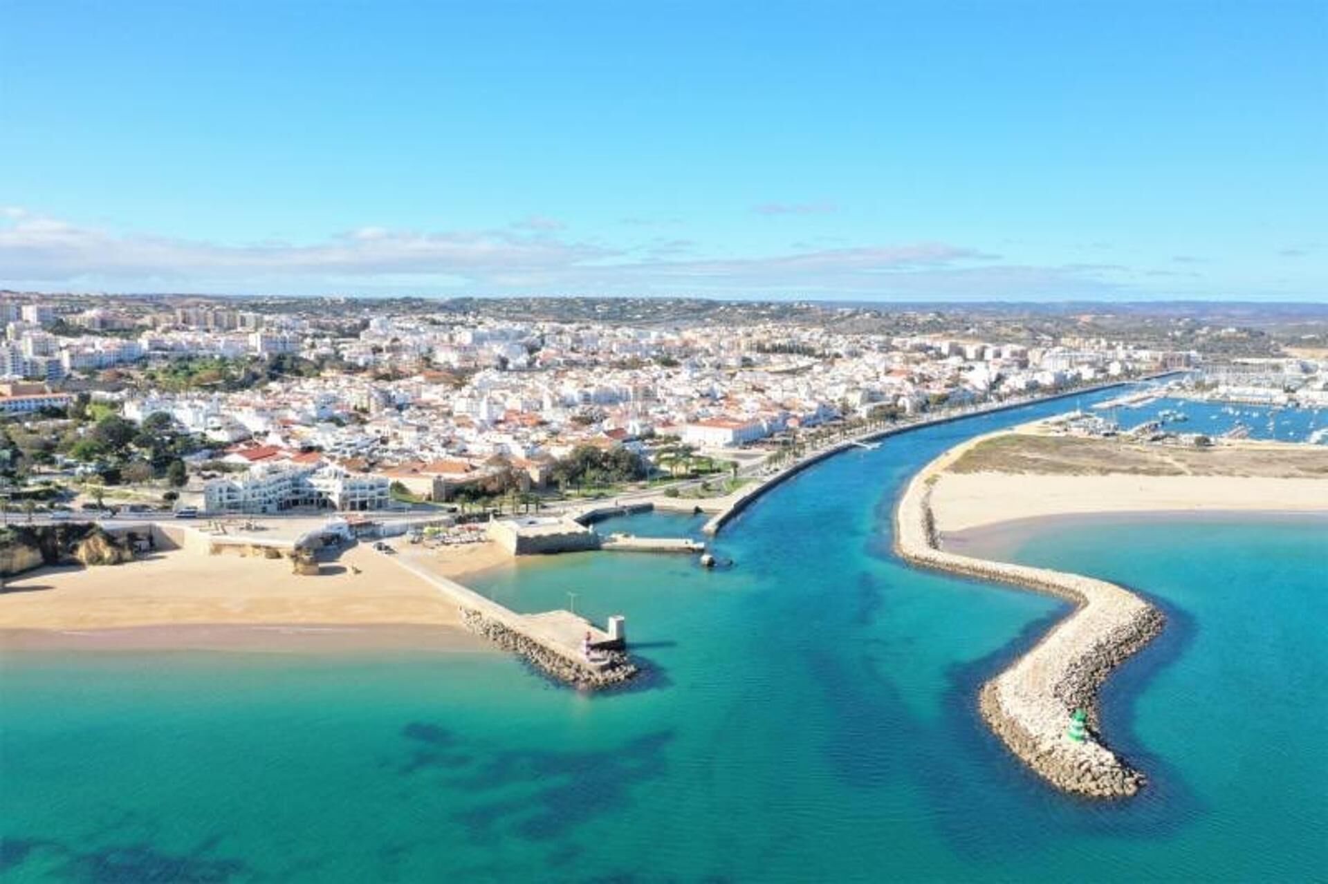 An aerial view of a coastal city with a prominent curved bay, a sandy beach, and a densely built urban area with white buildings.
