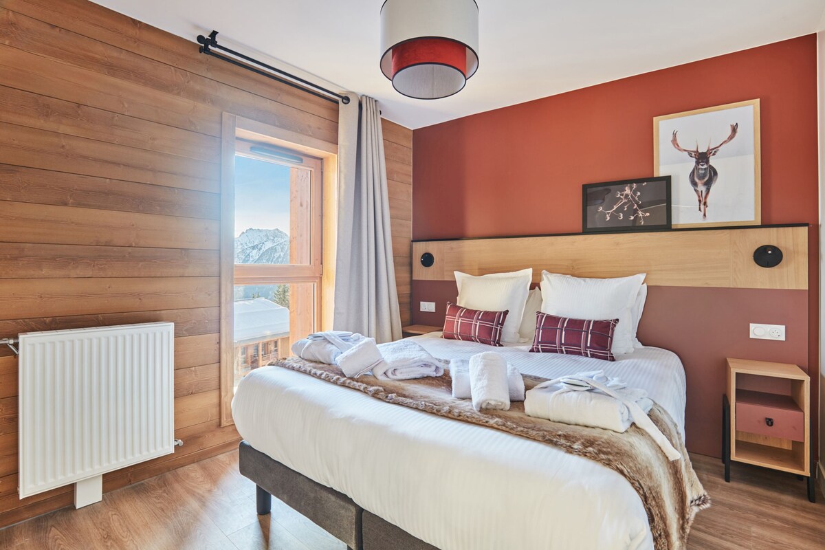Un dormitorio moderno con suelo de madera, una cama grande con ropa blanca, un cabecero con un cuadro colgado y vistas a las montañas a través de una ventana.