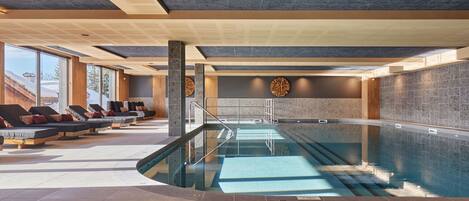 A modern indoor pool area with lounge chairs, a pool, and a view of the outdoors.