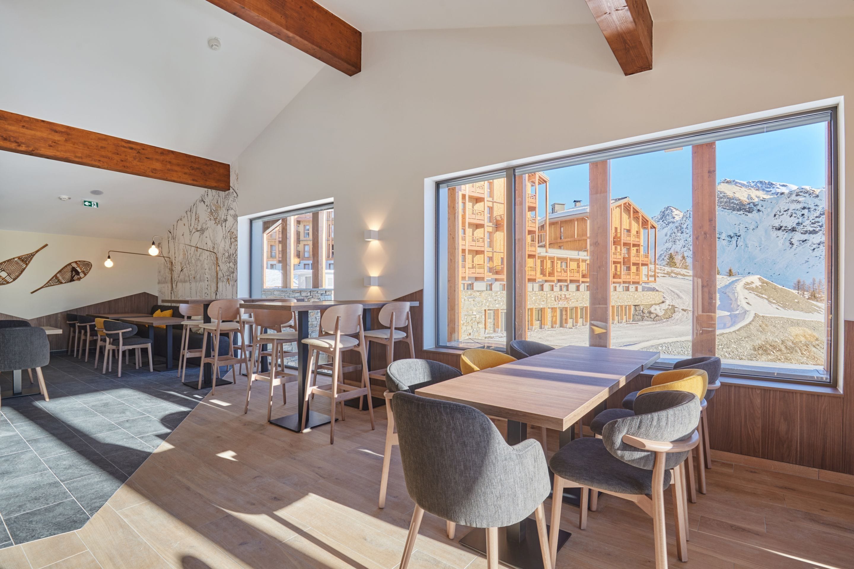 A modern dining area with wooden tables and chairs, large windows with a mountain view, and a stone wall with woven baskets.