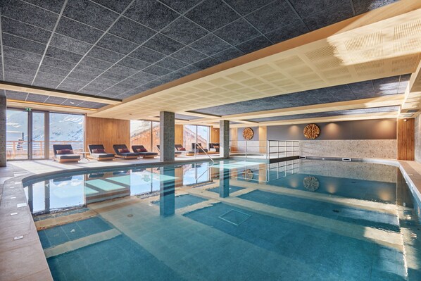 Una piscina interior con agua cristalina, tumbonas y vistas a montañas a través de amplios ventanales.