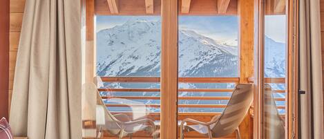 A room with a bed, two chairs, a window with a mountain view, and wooden walls.