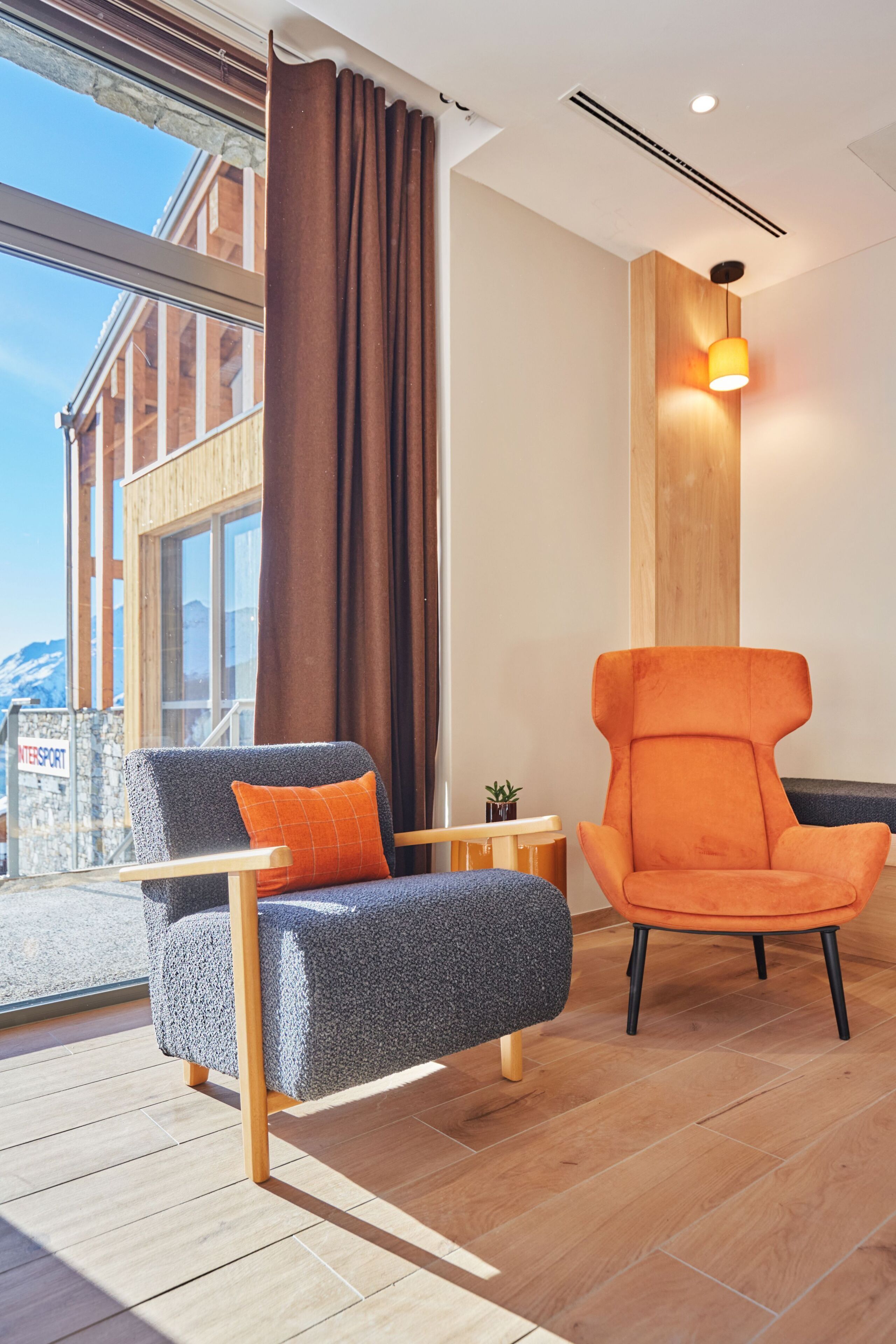 Ein modernes Wohnzimmer mit großem Fenster, Holzboden, grauem Sessel mit orangefarbenem Kissen und einem weiteren orangen Sessel.