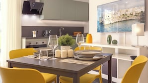A modern dining area with a table set for four, yellow chairs, a small potted plant, candles, and a painting of a coastal town. - Life70014  (Conversano)