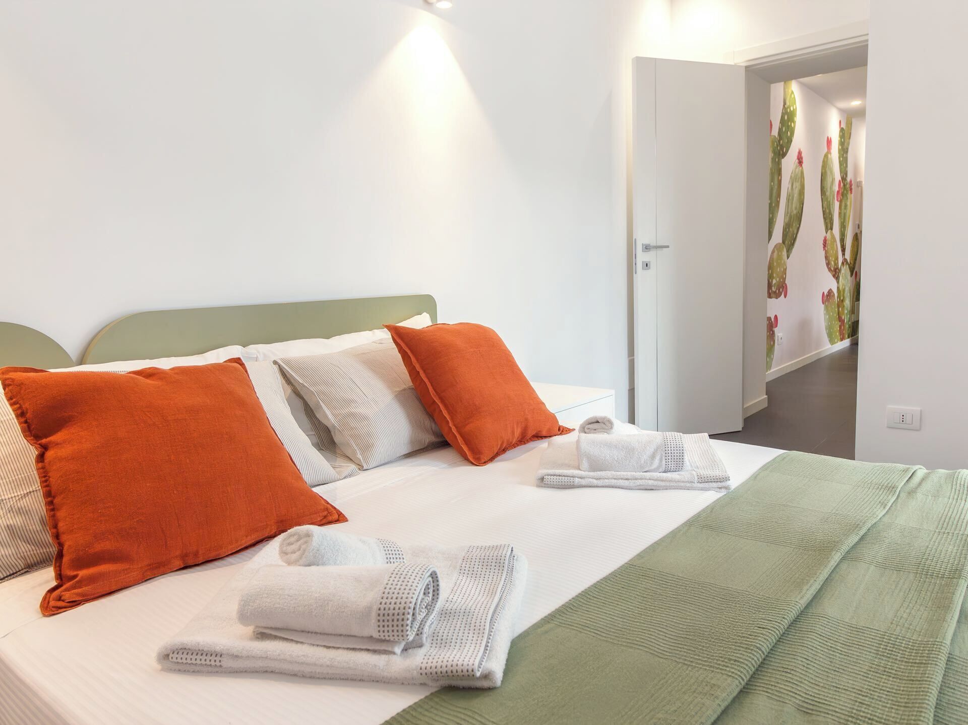 A neatly made bed with white and orange pillows, a green blanket, and rolled white towels.