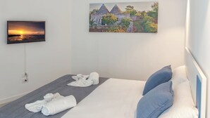 A neatly made bed with blue and white pillows, a grey blanket, and rolled towels. A painting of a building with a sunset is on the wall above the bed. - Life70014  (Conversano)