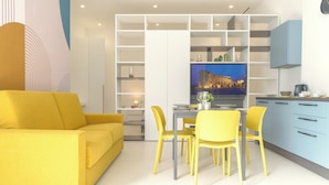 A modern living room with a yellow sofa and matching chairs, a dining area with a table and chairs, and a kitchen with blue cabinets and a television displaying a cityscape. - Life70014  (Conversano)
