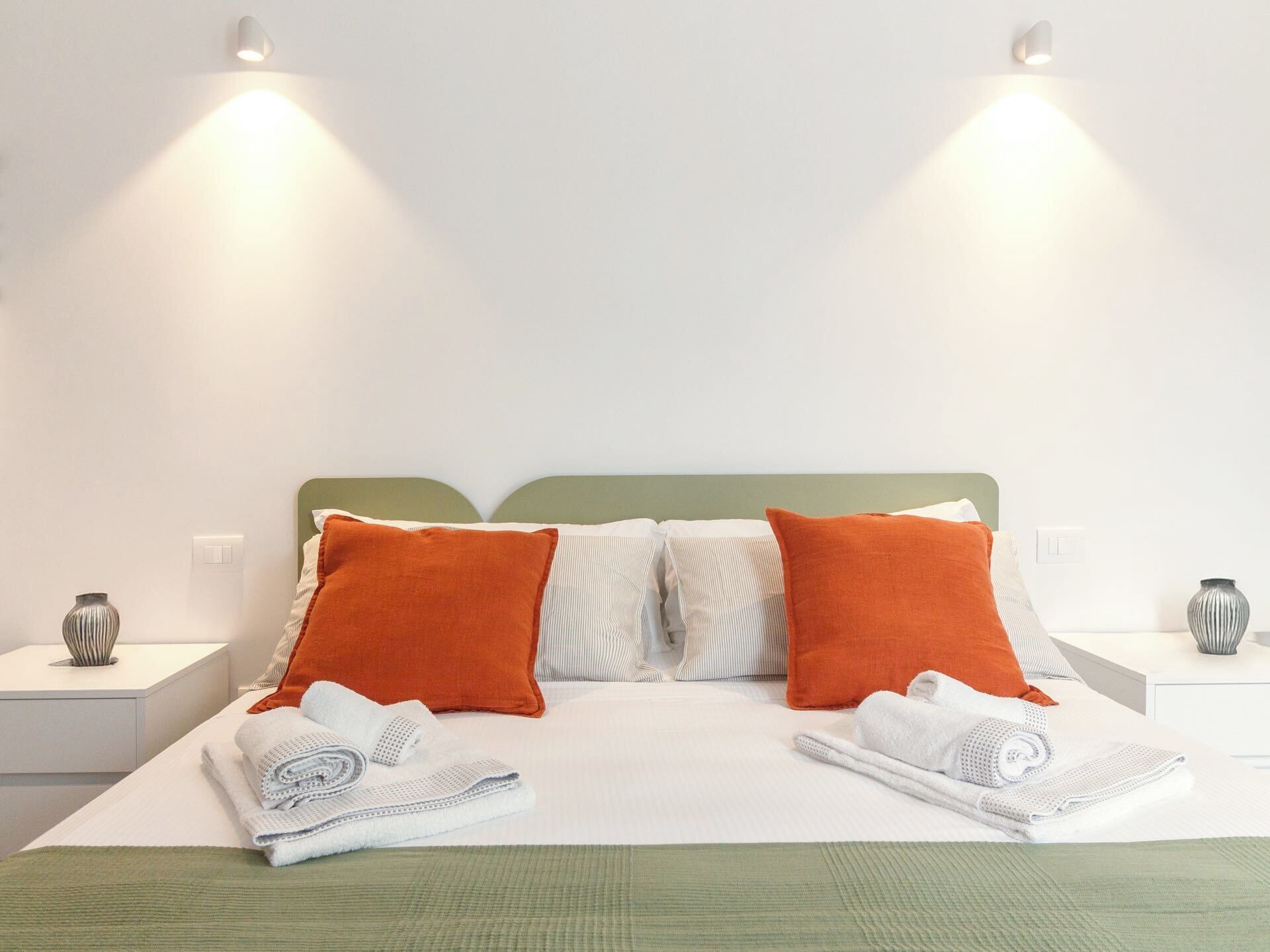 Una cama bien hecha con dos almohadas, un cabecero verde y dos mesitas de noche con jarrones.