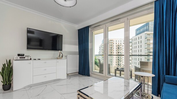 A modern living room with a flat-screen TV, white cabinetry, a marble coffee table, and a view of high-rise buildings through large windows. - Spektrum 2 by Grand Apartments (Gdansk)