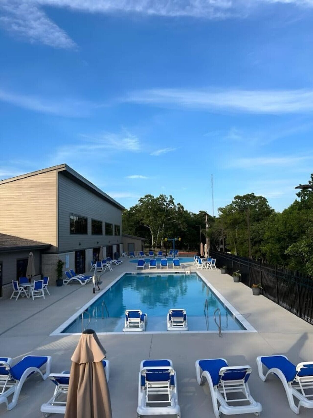A rectangular pool with blue lounge chairs and umbrellas, surrounded by a building with a flat roof and a fence.