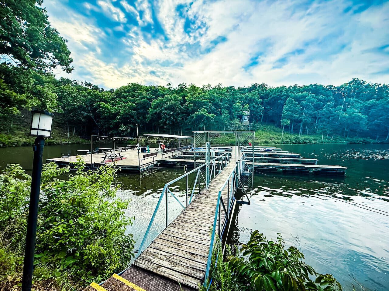 Eine hölzerne Pier erstreckt sich über ruhiges Wasser, umgeben von üppiger Vegetation und blauem Himmel.