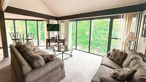 A spacious living room with two patterned sofas, a round coffee table, and a dining area with chairs and a table. Large windows provide a view of greenery outside. - Treetop Village Sunset Hollow #105-3BD/3BA/Pools/Grill/Dock/Deck-6PP (Village of Four Seasons)