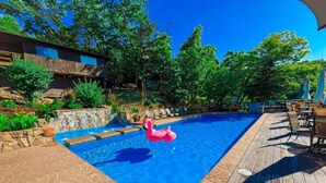 A swimming pool with a wooden deck, surrounded by greenery and a stone wall. - Treetop Village Sunset Hollow #105-3BD/3BA/Pools/Grill/Dock/Deck-6PP (Village of Four Seasons)