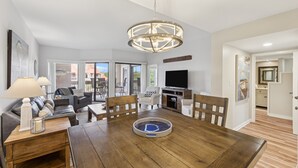 Ein geräumiges Wohnzimmer mit einem Holzesstisch, einer Chandelüster und einem Fernseher. Es gibt Sofas, Sessel und einen Couchtisch.