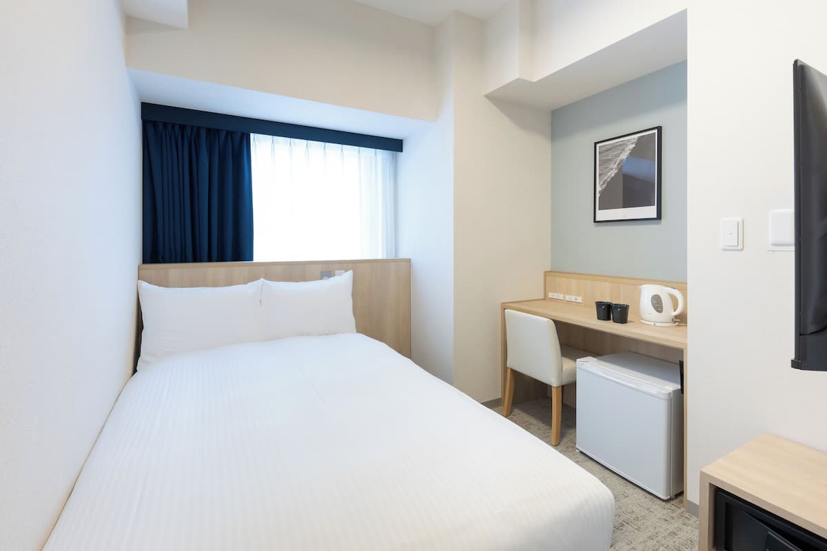 Hotelkamer met een bed, bureau, stoel, kleine koelkast en raam met gordijnen.