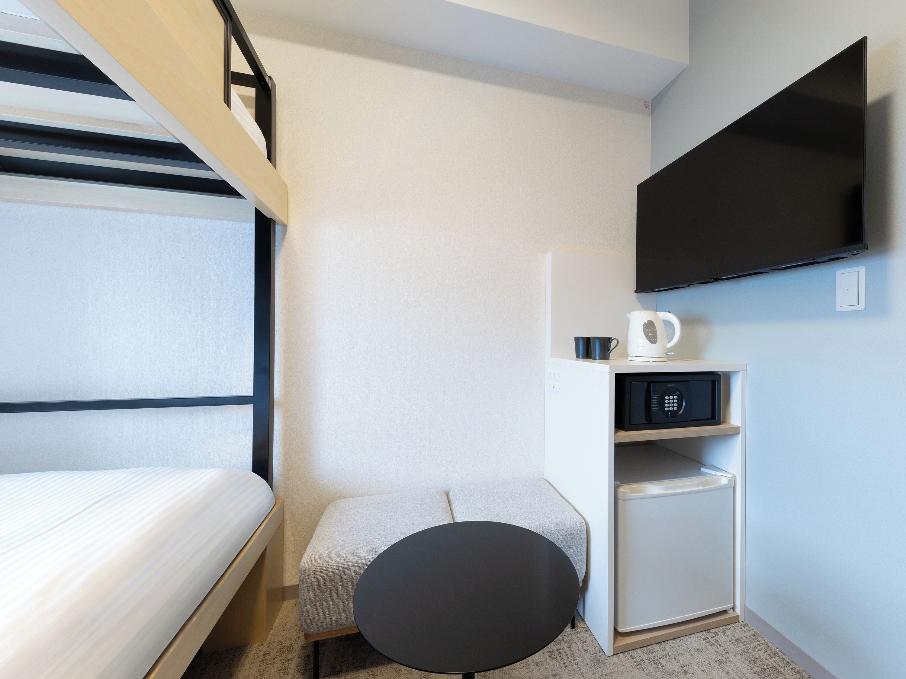 A compact hotel room with a bed, a small round table, a microwave, a kettle, and a TV mounted on the wall.