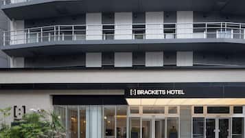 Un hotel moderno con fachada de vidrio, un cartel negro que dice "Brackets Hotel" y un área de entrada ajardinada.
