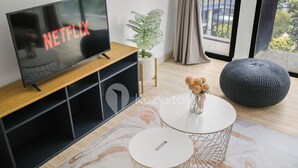 A modern living room with a flat-screen TV on a wooden stand, a potted plant, a round grey ottoman, and a small table with a vase of flowers. - Marmo by Kozystay - Kuningan (Jakarta)