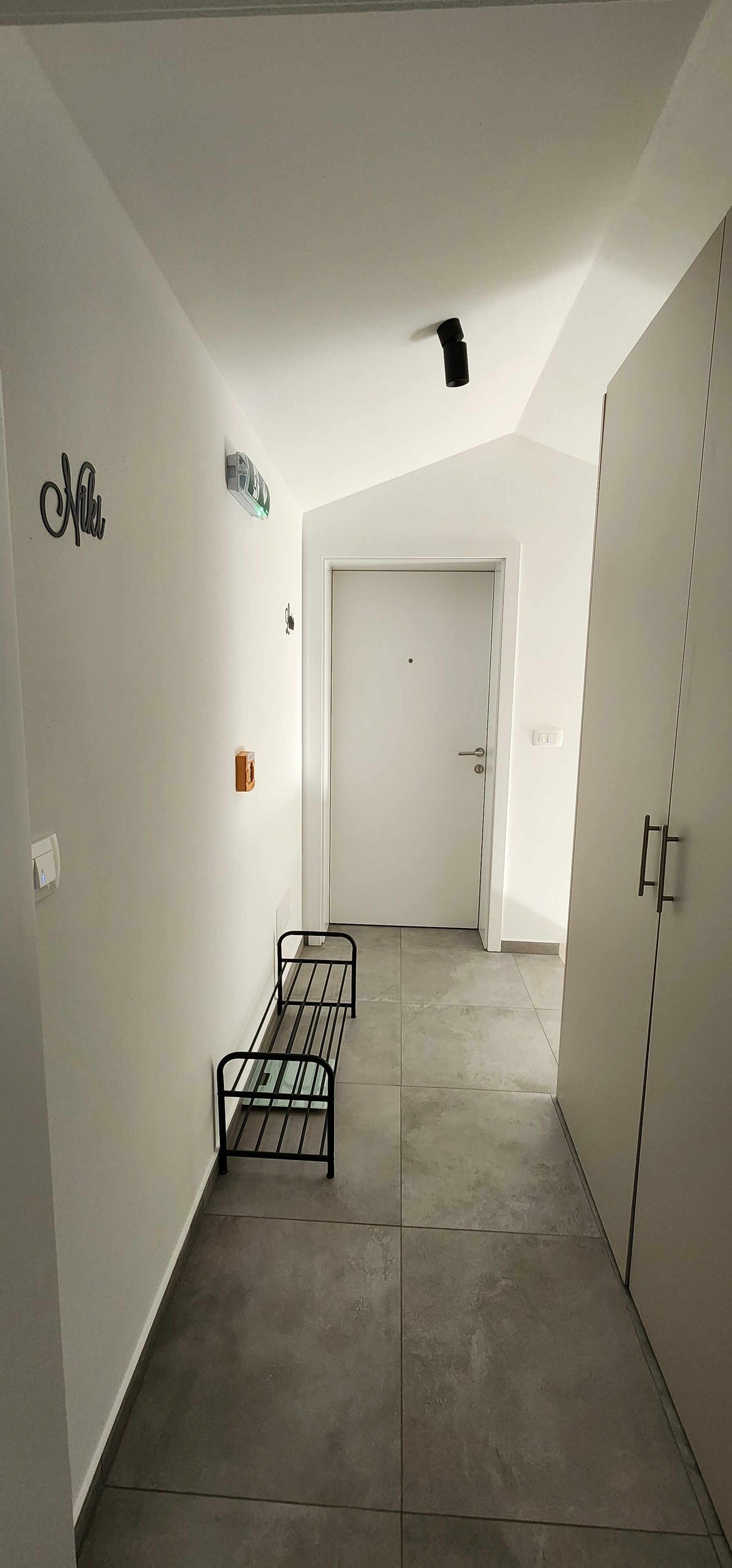 A narrow hallway with a white door at the end, a small shelf with a bottle, and a metal bench.