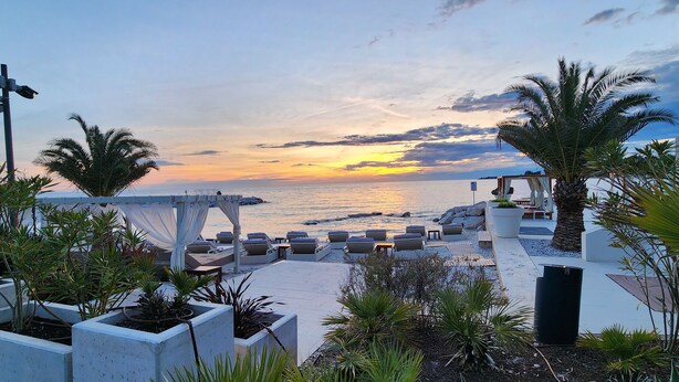 A beachside lounge area with seating and a pergola, overlooking the sea during sunset.
