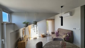 A modern living room with a sofa, coffee table, and hanging plants. There is a window with blinds, a fireplace, and a TV on a wooden stand.
