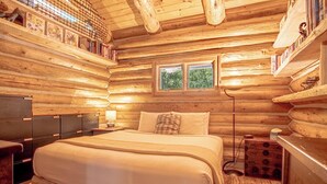 Ein Schlafzimmer in einer Blockhütte mit einem großen Bett, einem Nachttisch, einer Kommode und einem Fenster mit Blick auf Bäume.