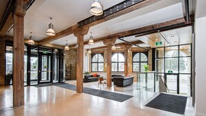 A spacious lobby with high ceilings, exposed wooden beams, large windows, and a seating area with a sofa and chairs. - Bradshaw Lofts: The Tapestry (Stratford)