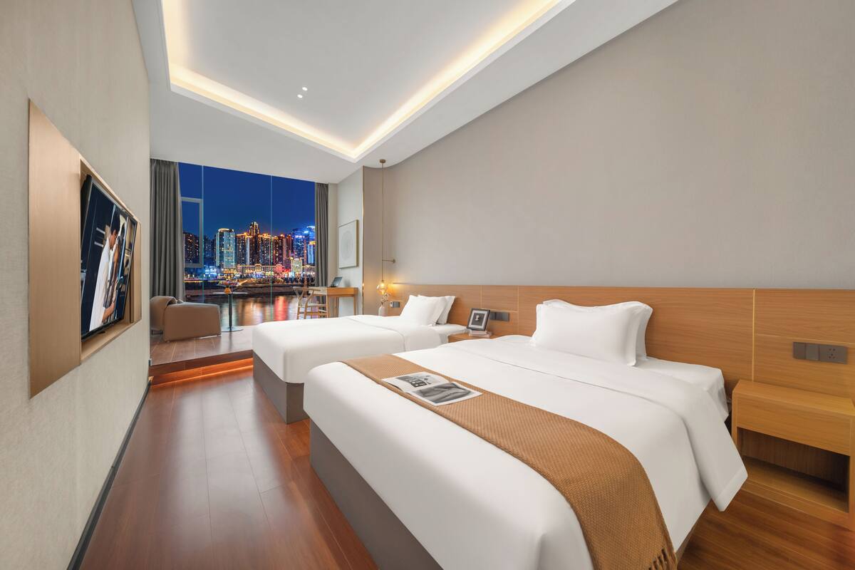 A modern hotel room with two beds, a city view, and a dining area with a table and chairs.