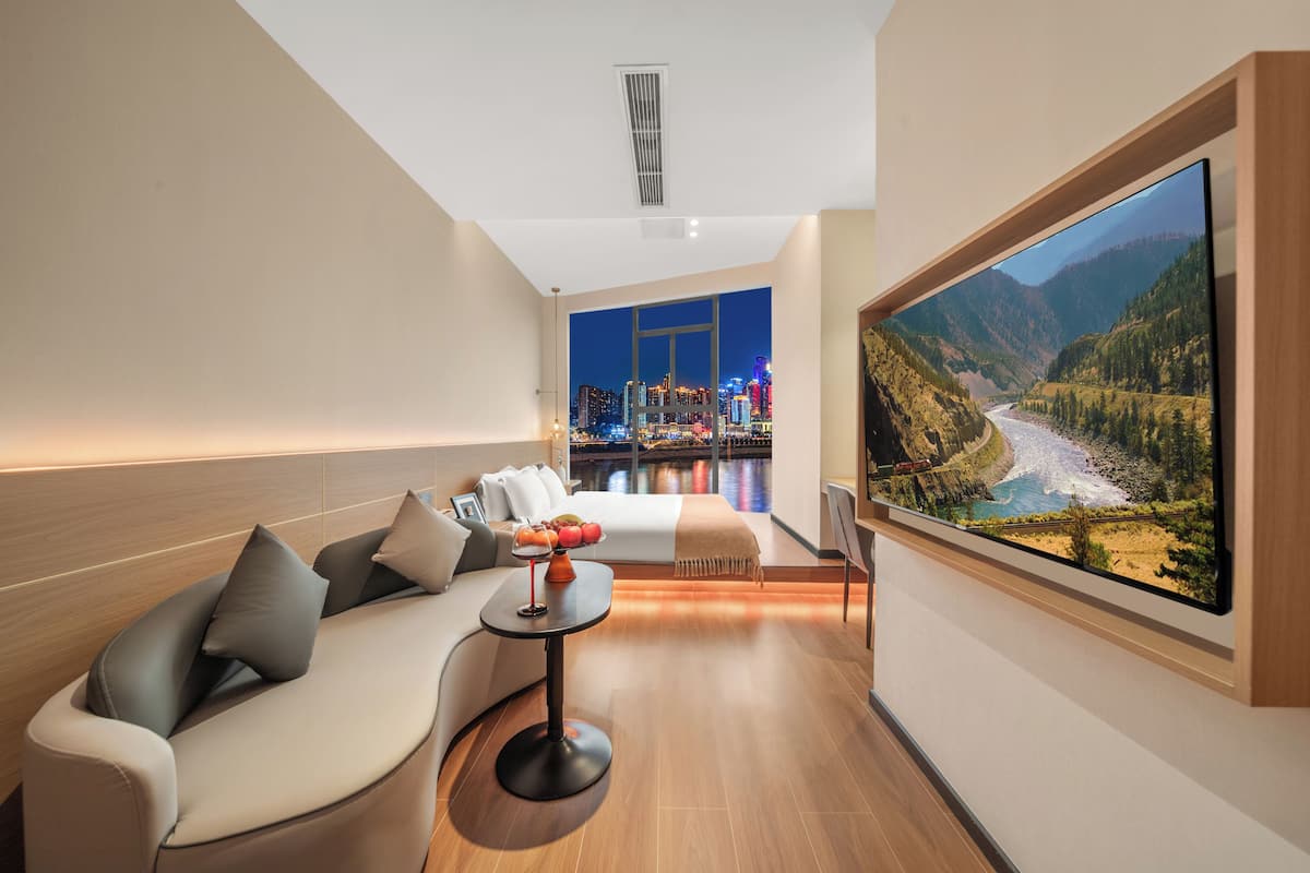 A modern hotel room with a large TV displaying a scenic mountain view, a sofa, a small table with fruit, and a cityscape view through the window.