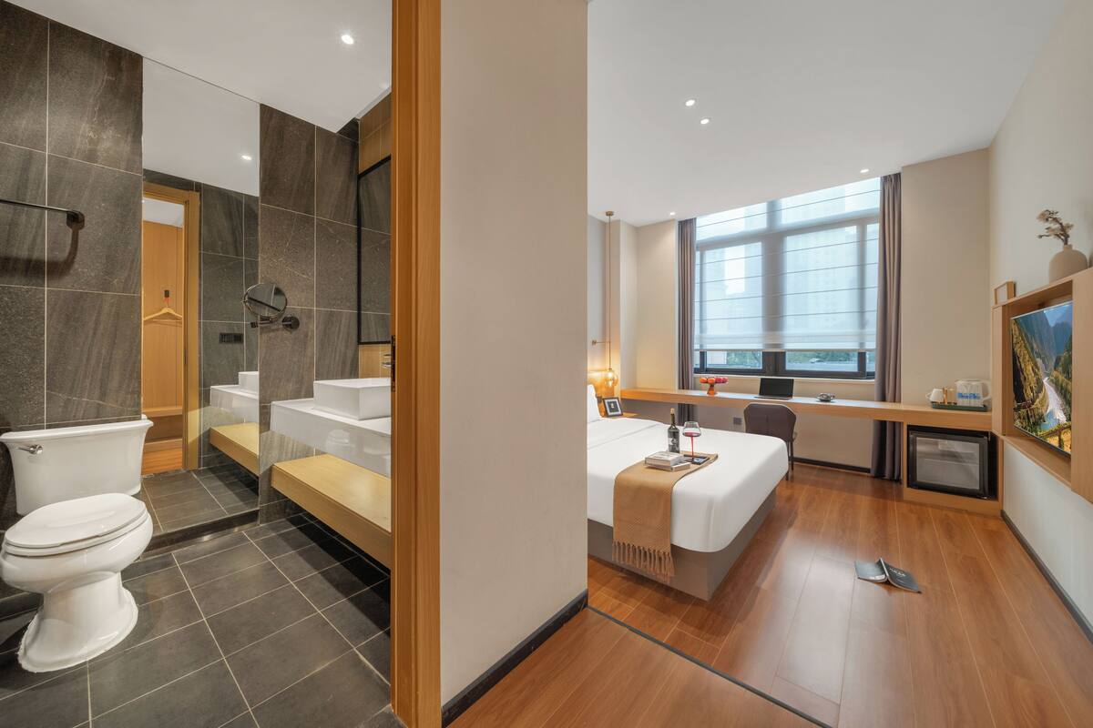 A modern hotel bathroom with a toilet, shower, and sink. There is a bed with a bedside table and a window with a view.