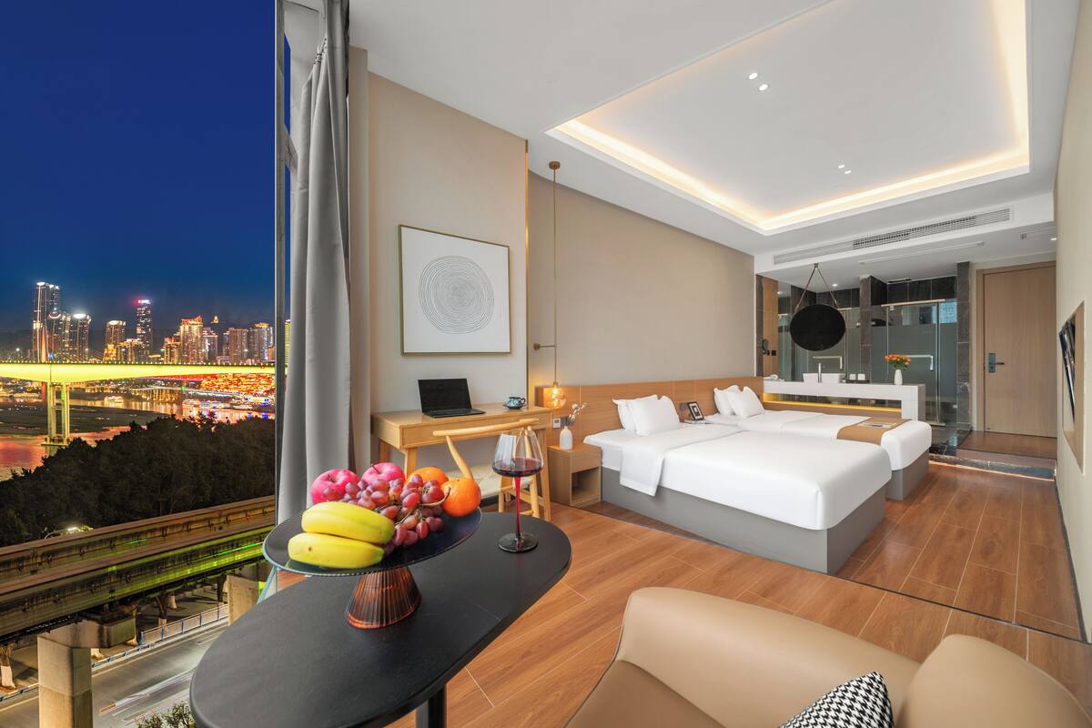 A modern hotel room with a large bed, a desk with a laptop, a dining table with fruit, and a city view.