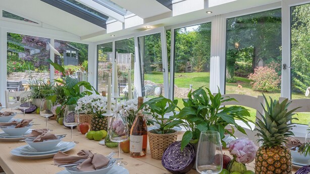 Une table à manger dressée pour un repas, avec vue sur un jardin grâce à de grandes fenêtres.
