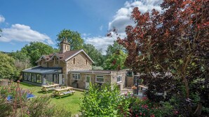 Une maison en pierre avec une véranda, une terrasse avec des meubles d’extérieur et un jardin agrémenté de diverses plantes et fleurs.