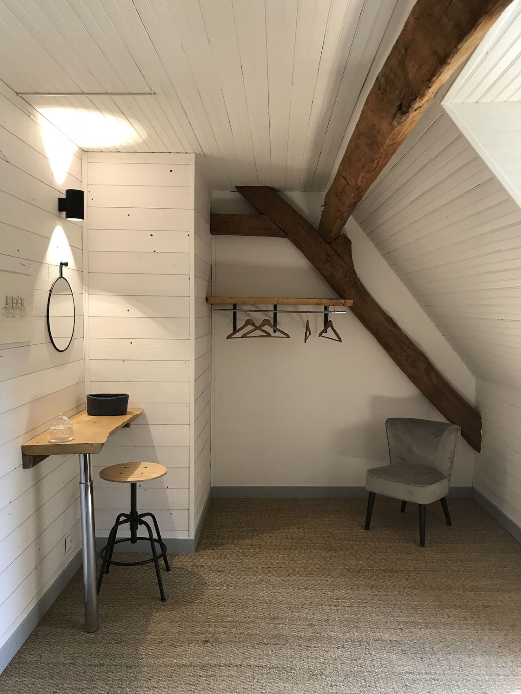 A small, cozy attic room with a slanted ceiling, a wooden table with a stool, a chair, a coat rack with hangers, and a wall-mounted light fixture.