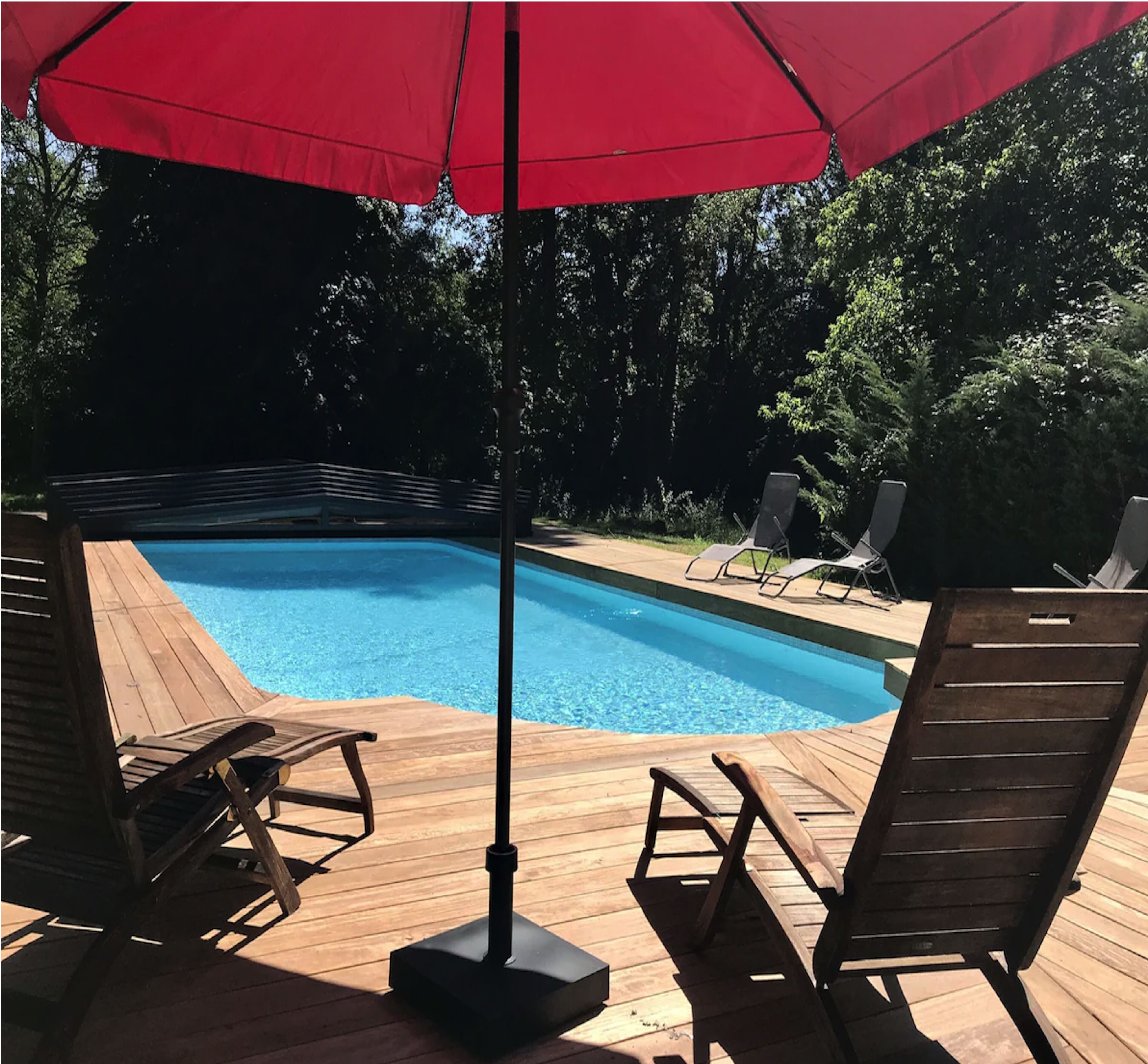A swimming pool with a wooden deck, surrounded by lounge chairs and a red umbrella.