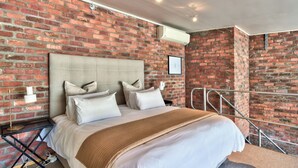 A bedroom with a brick wall, a bed with white bedding, a headboard, and a bedside table with a lamp. - West Side Studios Mountain View Loft (Cape Town)