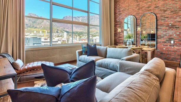 A modern living room with a large window, a brick wall, and a view of a cityscape. - West Side Studios Mountain View Loft (Cape Town)