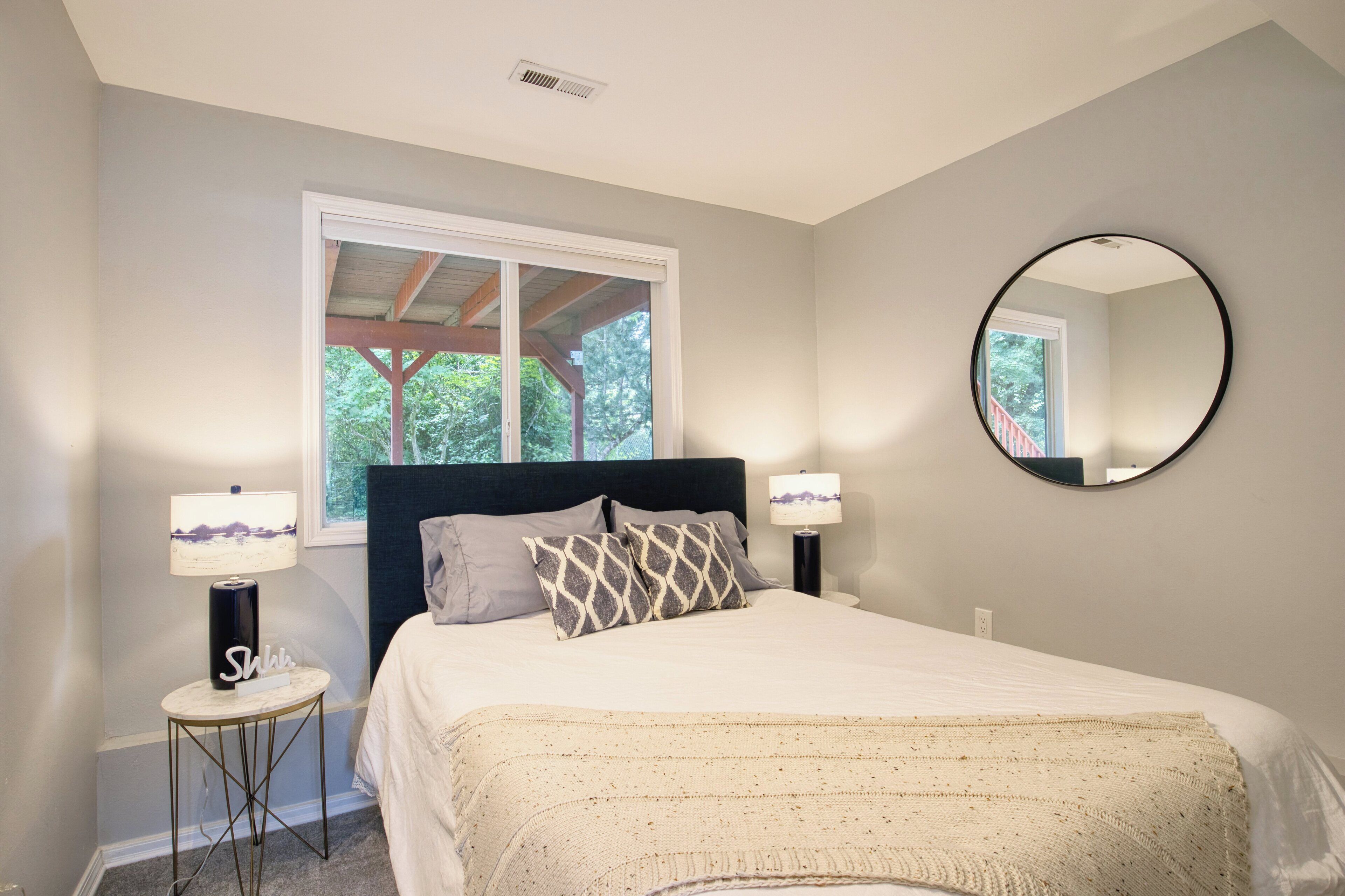 A bedroom with a bed, two bedside lamps, a round mirror, and a window with a view of trees.