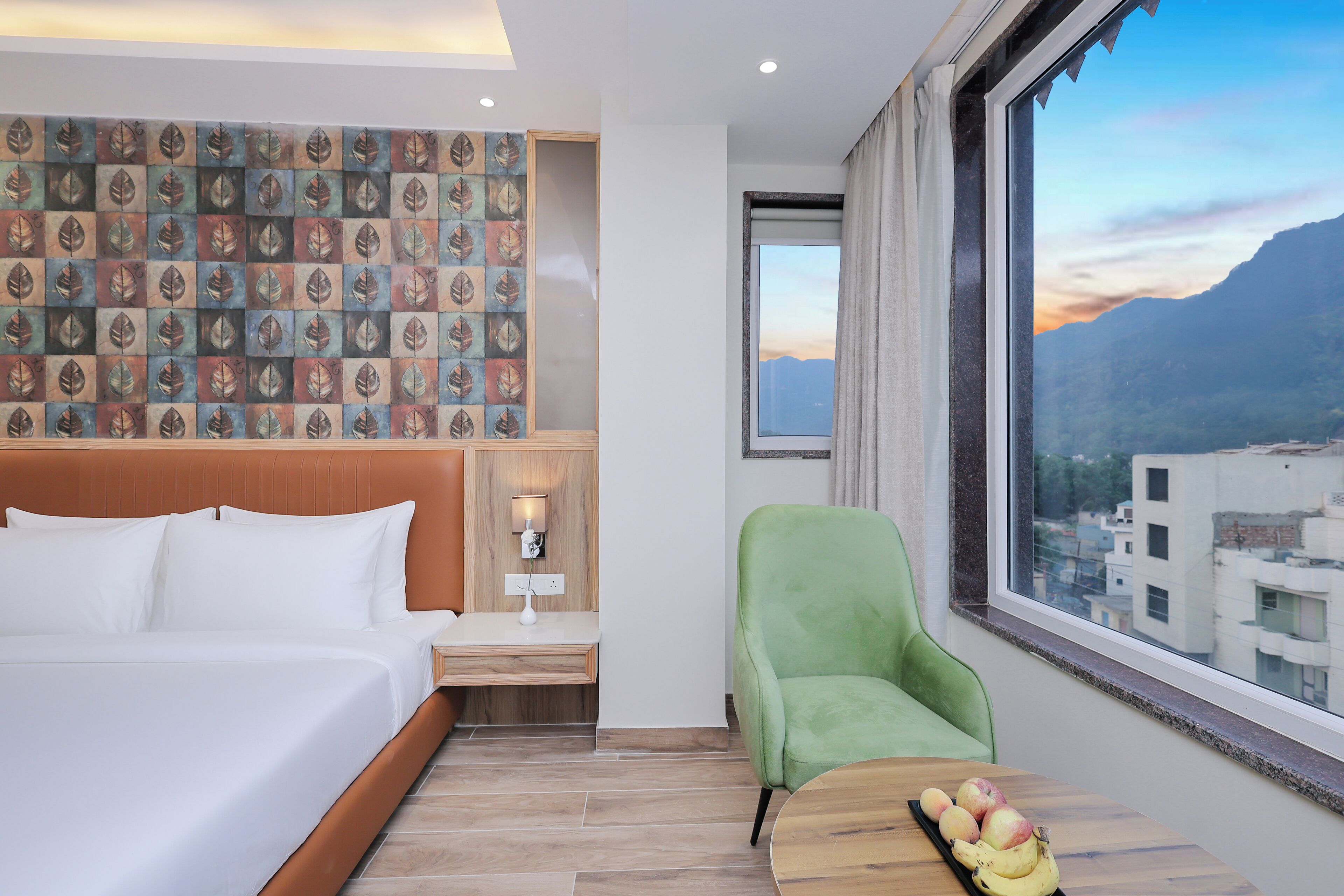 A hotel room with a bed, a desk, a chair, and a view of a cityscape through a large window.