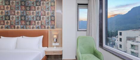 A hotel room with a bed, a desk, a chair, and a view of a cityscape through a large window.
