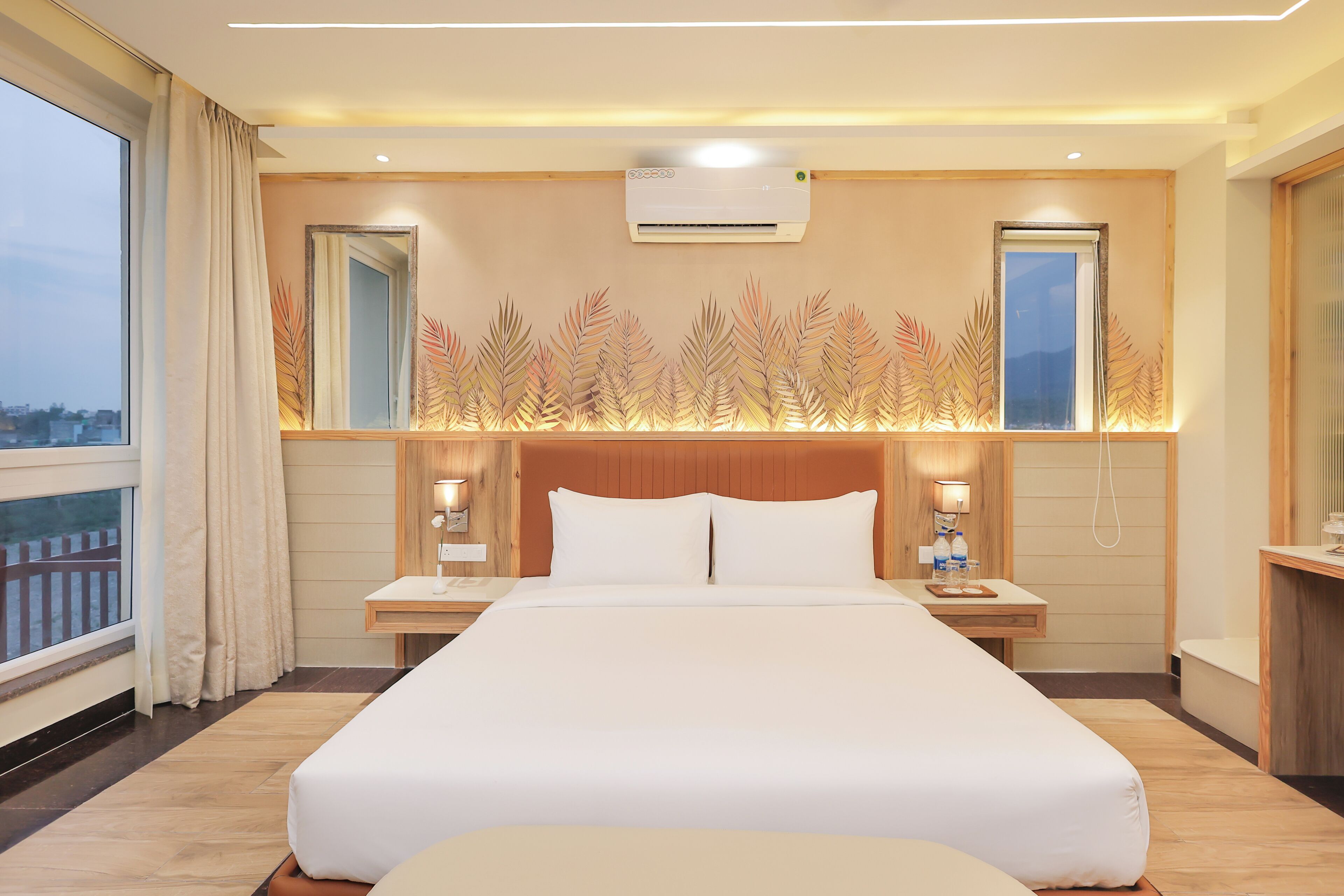 A hotel room with a large bed, wooden headboard, bedside lamps, a nightstand, a window with curtains, and a wall-mounted air conditioner.