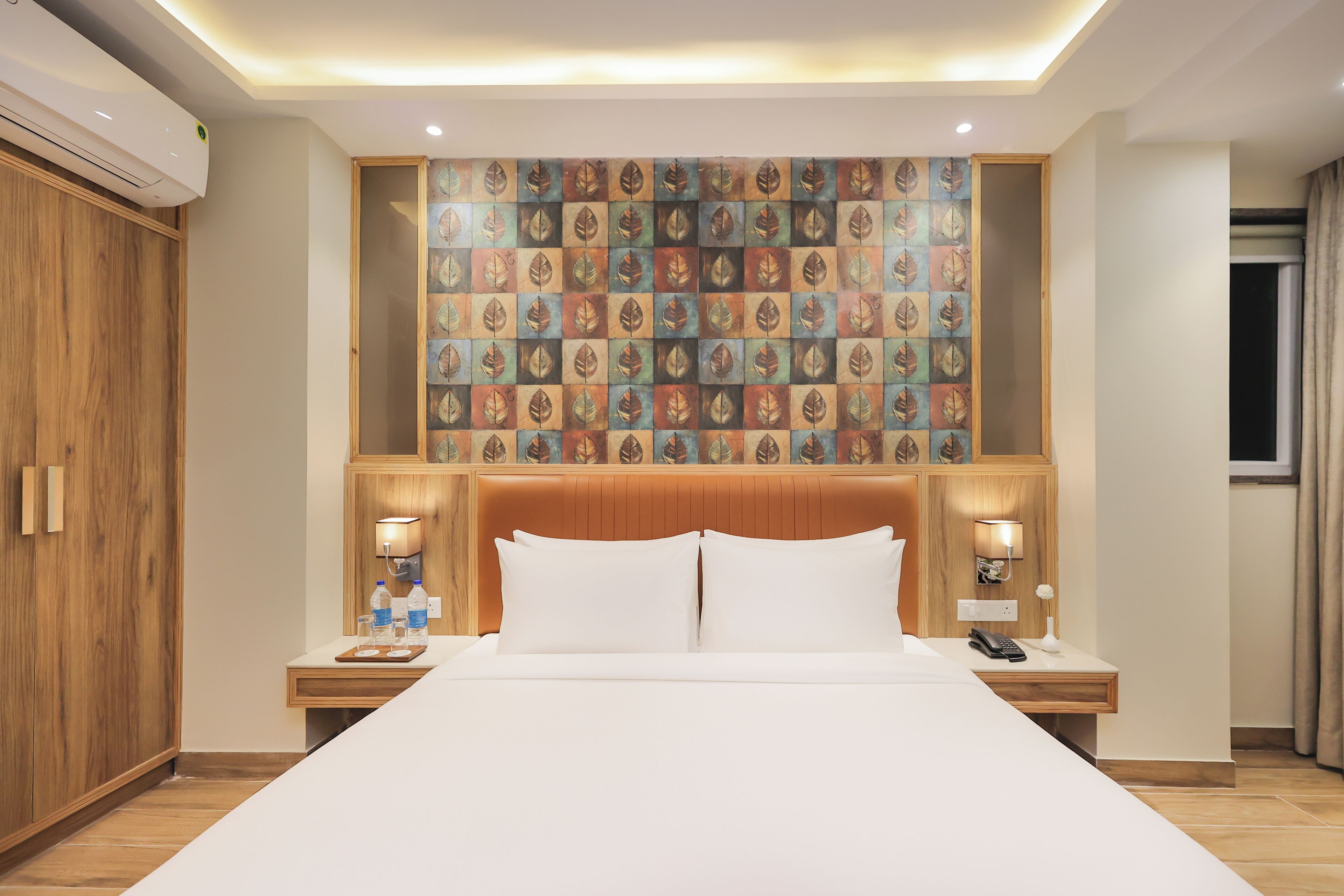 Une chambre d’hôtel avec un grand lit, une tête de lit en bois, deux tables de chevet avec des lampes et un mur décoratif orné d’un motif de feuilles.