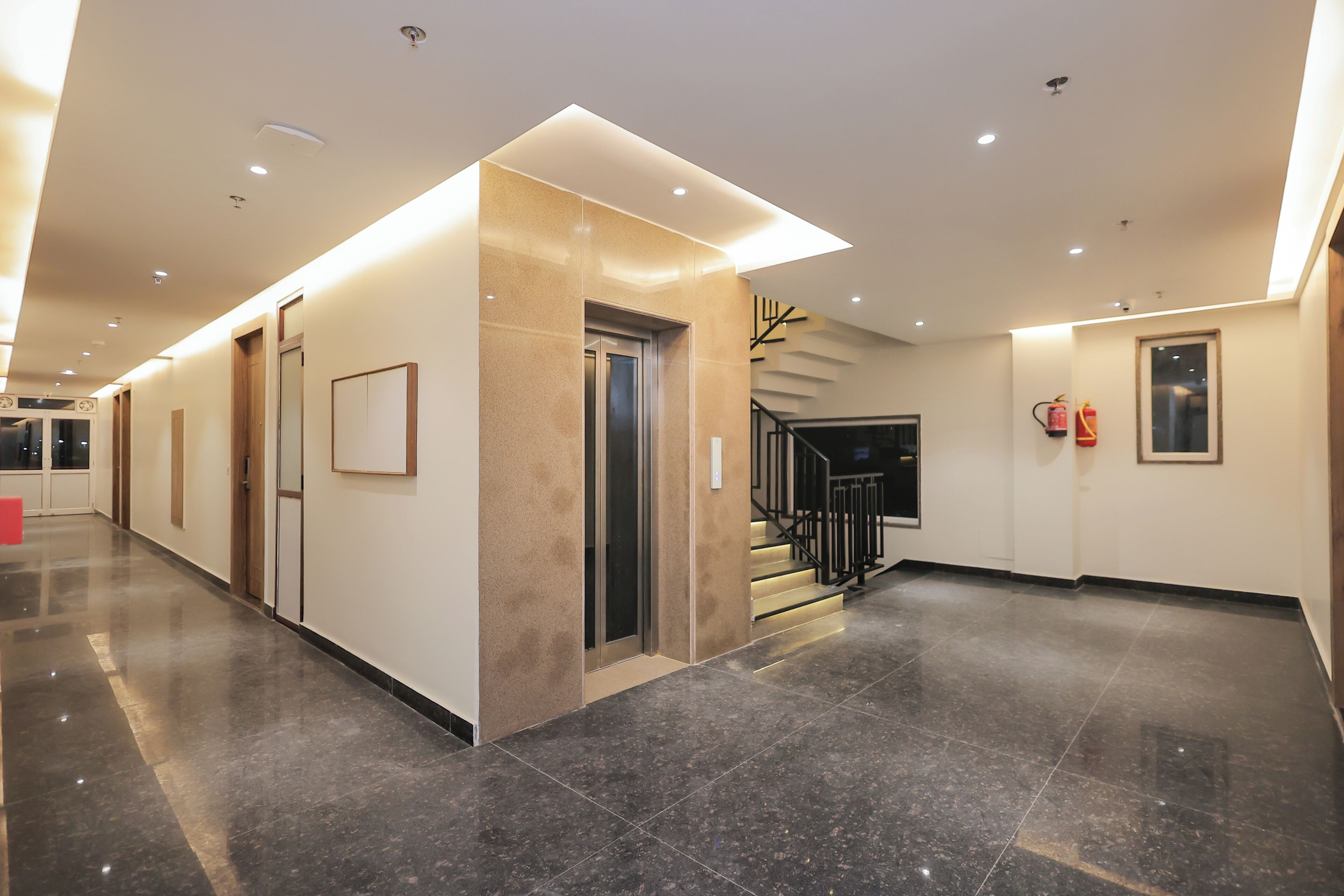 A spacious hotel lobby with a modern design, featuring a marble floor, a central elevator, and a staircase with black railings.