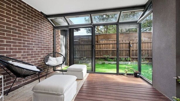 A modern patio with a brick wall, a wooden deck, a glass door leading to a backyard, and two white cushioned seating areas.